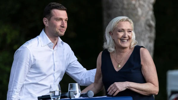 Marine Le Pen et Jordan Bardella souriants derrière un pupitre avec microphones et verres d'eau lors d'une apparition publique en extérieur