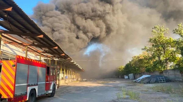 Camion de pompiers rouge stationné sur un chemin de terre près de l'entrepôt de la Croix-Rouge à Kherson d'où s'élève un épais panache de fumée noire après une attaque