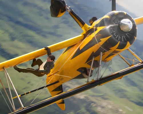 Tom Cruise suspendu à l'aile d'un biplan jaune volant à l'envers au-dessus d'un paysage verdoyant lors du tournage de la suite de Mission: Impossible