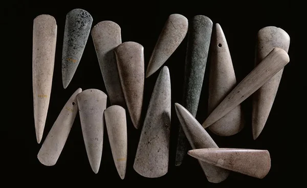 Dépôt de haches polies en pierre de différentes tailles et couleurs (grise à verte), disposées sur fond noir. Certains objets présentent des trous de perforation.
