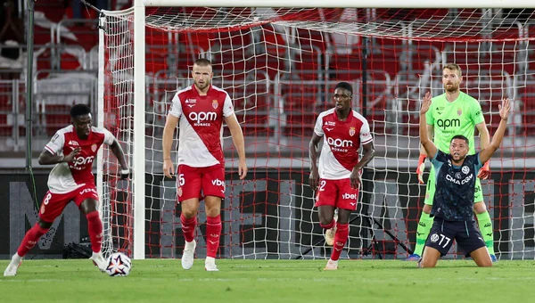 Un joueur de l'OM en tenue bleue agenouillé sur le terrain avec les bras levés, tandis que des joueurs de Monaco en rouge et blanc et le gardien en vert se tiennent près du but lors du match Ligue 1 où Monaco a battu l'OM 2-0.
