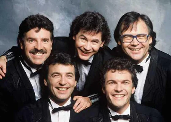 Portrait de groupe des cinq membres des Musclés en smokings noirs avec nœuds papillon, souriant devant un fond gris marbré.