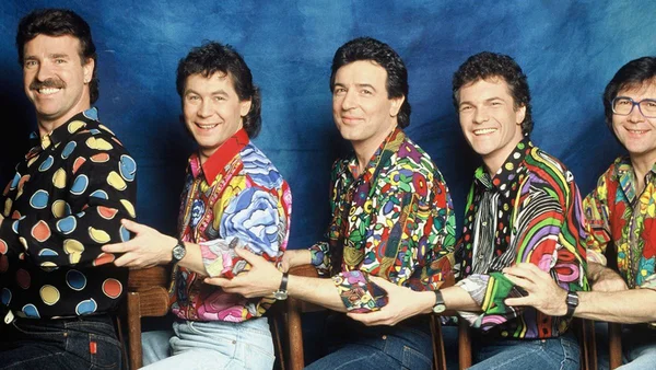 Framboisier, chanteur des Musclés du Club Dorothée, avec les autres membres du groupe, assis en rang sur fond bleu, vêtus de chemises à motifs colorés et jeans, chacun posant la main sur l'épaule du voisin.