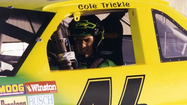 Un pilote de course portant un casque et des lunettes assis dans une voiture de course jaune, avec le nom 'Cole Trickle' écrit au-dessus de la fenêtre.