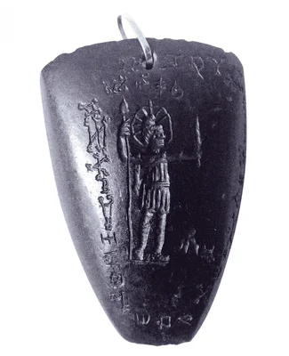 Photographie en noir et blanc d'un amulette en plomb triangulaire en relief, représentant une figure debout tenant une lance, entourée d'inscriptions.