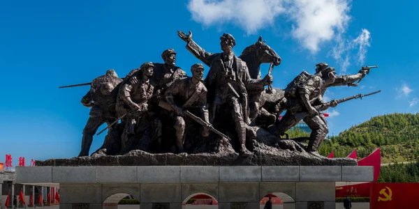 Grande sculpture en bronze représentant un groupe de soldats en poses dynamiques, avec des drapeaux rouges dont un à la faucille et au marteau, sur fond de ciel bleu et de collines verdoyantes, évoquant la Longue Marche.