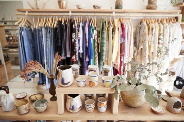 Une exposition commerciale avec une table en bois ornée de poteries céamiques faites main, de fleurs sèches et d'une plante en pot, avec un présentoir de vêtements colorés en arrière-plan
