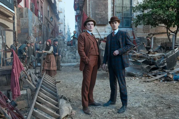 Hero Fiennes Tiffin et un autre homme en costumes et chapeaux des années 1920 dans une rue jonchée de débris, avec une foule et une barricade en arrière-plan.