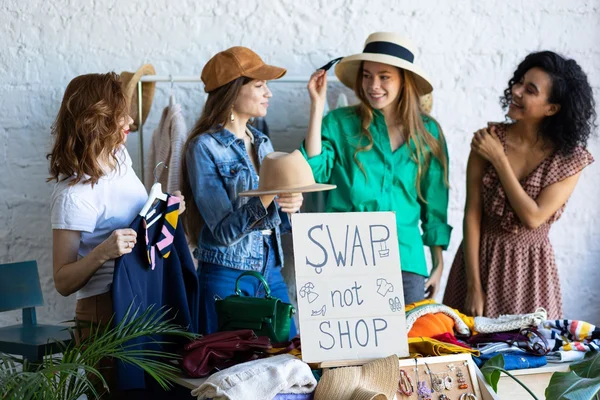 Quatre femmes entouées de vêtements et d'accessoires sur une table, souriant et interagissant, avec un panneau 'SWAP not SHOP' en premier plan, lors d'un échange de vêtements entre amis