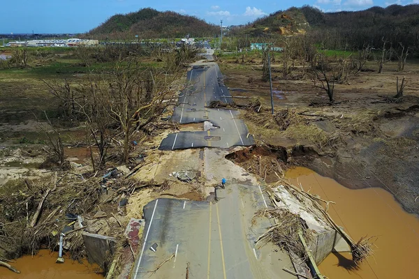 Vue aérienne d'une route en asphalte sévèrement endommagée et fragmentée à Porto Rico après un désastre, avec des débris et de l'eau boueuse.
