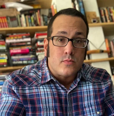 L'auteur Gabino Iglesias avec des cheveux courts foncés, des lunetes et une chemise à carreaux bleue et rouge, regardant la caméra devant des étagères de livres.