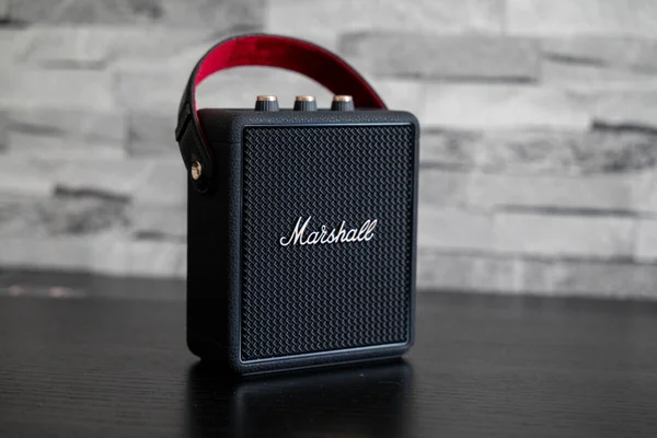 Enceinte bluetooth portable Marshall Stockwell III noire avec sa sangle de transport rouge, posée sur une surface sombre devant un mur de briques.