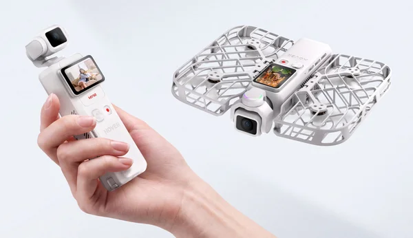Un drone blanc HoverAir Versa avec cage de protection et caméra, volant près d'une main tenant une manette blanche avec petit écran, sur un fond uni.