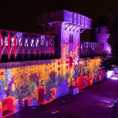 Projection lumineuse colorée sur une tour et un rempart du château de Carcassonne lors de 'Remparts et Lumières', révélant les détails architecturaux et une hourd en bois.