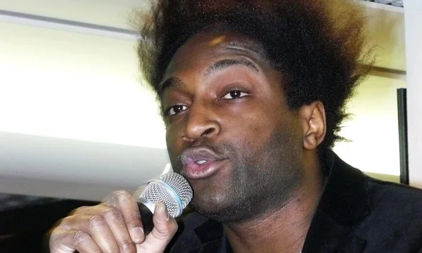 Portrait en gros plan de Kamini, avec sa grande afro, chantant ou parlant en main un micro lors d'un concert ou d'un tournage