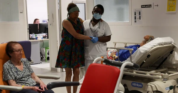 Une soignante en uniforme blanc et masque se tient à côté d'une femme en robe à fleurs, tandis qu'une femme âgée est assise sur une chaise orange et qu'un autre patient est allongé sur un lit d'hôpital en arrière-plan, illustrant la crise hospitalière lors de la canicule.