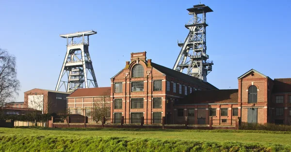Bâtiment industriel en briques rouges avec deux chevalements en acier, site minier classé au patrimoine mondial de l'UNESCO dans le bassin minier du Nord-Pas-de-Calais, sous un ciel dégagé et un champ vert.