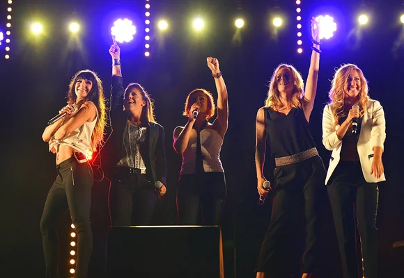 Les cinq membres du girl group L5 en pleine performance sur scène, brandissant des micros sous des projecteurs éclatants.