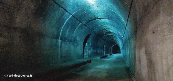 Galerie souterraine voûtée en béton, éclairée de manière tamisée avec une teinte bleuâtre, présentant une ouverture sombre sur la gauche, typique des lieux souterrains de la région.