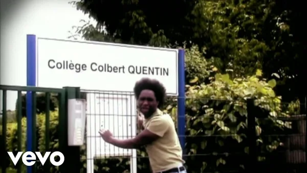 Kamini, chanteur français afro et en t-shirt jaune, debout devant le panneau du Collège Colbert à Quentin