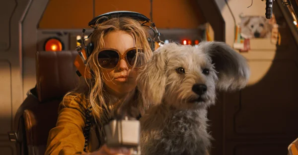 Milly Alcock dans le rôle de Supergirl 'punk rock', assise dans un cockpit avec des lunettes de soleil et un casque, accompagnée d'un petit chien blanc.