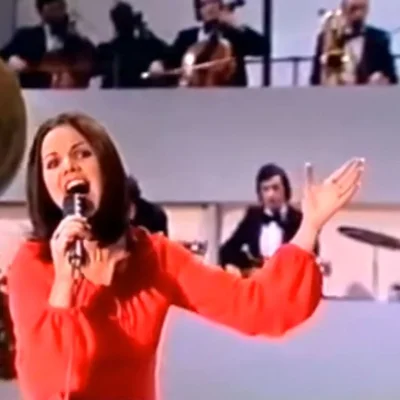 Une femme aux cheveux foncés vêtue d'un haut rouge chantant dans un microphone lors d'une performance Eurovision, accompagnée de musiciens en arrière-plan.
