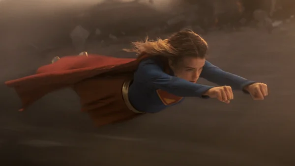 Milly Alcock en Supergirl volant horizontalement dans un environnement poussiéreux et rempli de débris.