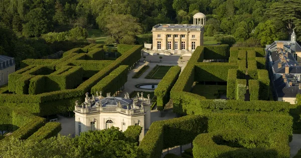 Vue aérienne du Petit Trianon au Château de Versailles, avec des jardins formels, des labyrinthes de haies, des pavillons classiques et un château central entouré de verdure.