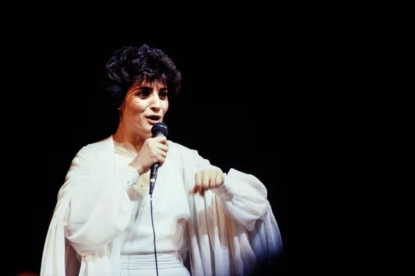Linda de Suza, chanteuse populaire des années 80, avec des cheveux courts et sombres, portant une tenue blanche, chantant dans un microphone sur fond noir.