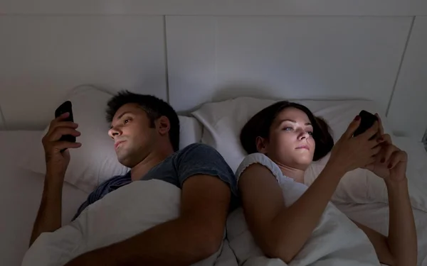 Couple allongé dans un lit, chacun regardant son écran de smartphone dans une pièce sombre, illustrant la routine et les difficultés de communication dans le couple.