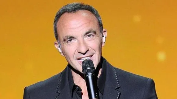 Nikos Aliagas en costume sombre chantant dans un micro sur fond jaune pendant un épisode de la Star Academy.