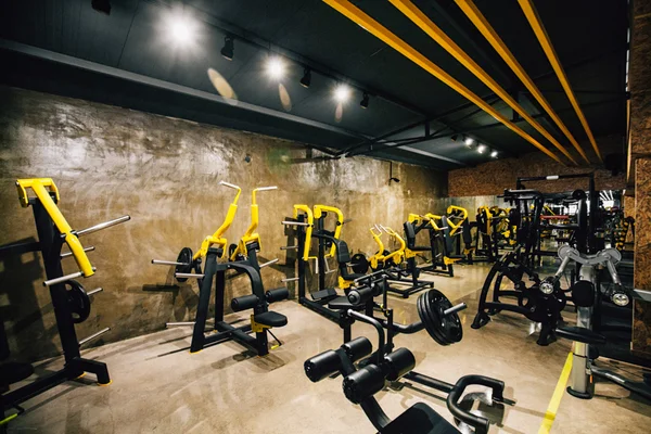 Intérieur d'une salle de sport moderne avec des machines de musculation noires et jaunes sur sol béton, éclairage directionnel et poutres décoratives au plafond