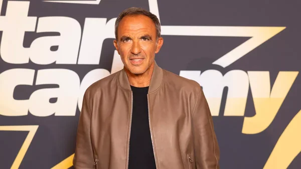 Nikos Aliagas en veste en cuir marron devant le logo de la Star Academy, lors d'un événement de promotion de la nouvelle saison.