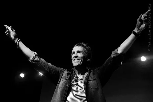 Jean-Patrick Capdevielle sur scène, bras levés, portant une chemise rayée sous une veste ouverte lors de son concert à Gien le 20 mai.