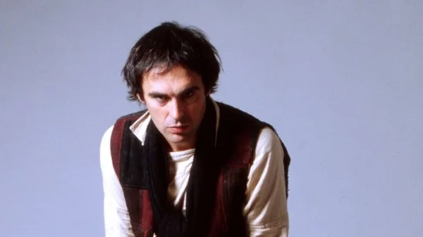 Portrait en studio de Jean-Patrick Capdevielle avec des cheveux sombres en désordre, une chemise blanche et un gilet sombre, sur fond bleu pâle.