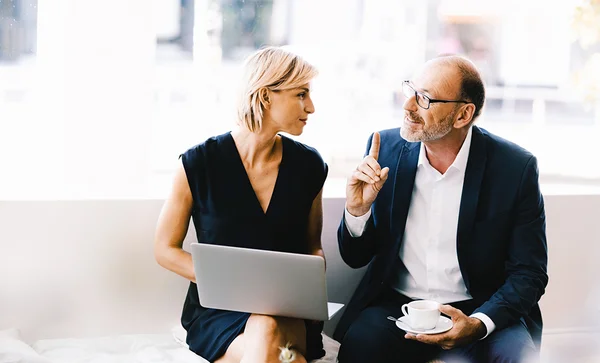 Réunion entre un conseiller professionnel et un client dans un bureau, où la femme tient un ordinateur portable sur ses genoux tandis que l'homme gesticule du doigt tout en tenant une tasse de café.