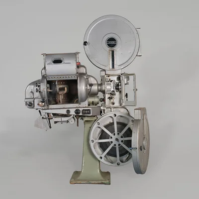 Projecteur de film 35 mm vintage argenté sur un pied vert pâle, avec deux grandes bobines et des mécanismes exposés, exposé en fond gris à la Cinémathèque française.
