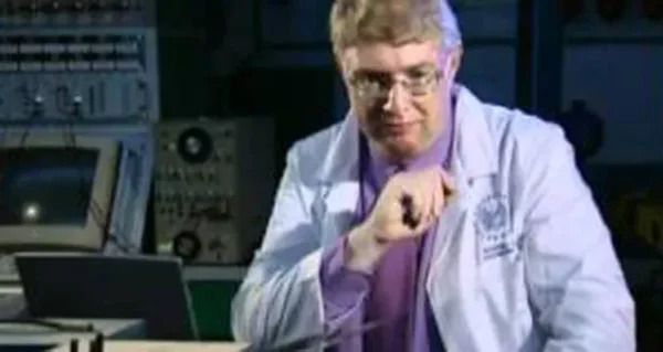 Vic Tandy, scientifique aux cheveux blonds portant des lunettes et une blouse de laboratoire blanche par-dessus une chemise violette, dans un laboratoire avec du matériel électronique.