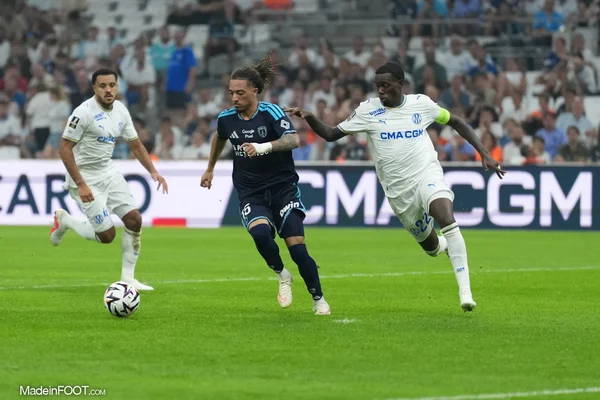 Action de jeu lors du match Ligue 1 entre l'OM et le Paris FC, montrant trois joueurs en compétition pour le ballon sur un terrain vert avec spectateurs et panneaux publicitaires en arrière-plan.