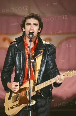 Jean-Patrick Capdevielle, en 1981, en veste en cuir noir et écharpe rouge, chantant dans un microphone tout en jouant de la guitare électrique sur scène.