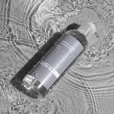 Photographie en noir et blanc d'une bouteille de lubrifiant intime longue durée YESforLOV 'Yes Get Love' à 100% silicone dans de l'eau ondulante.