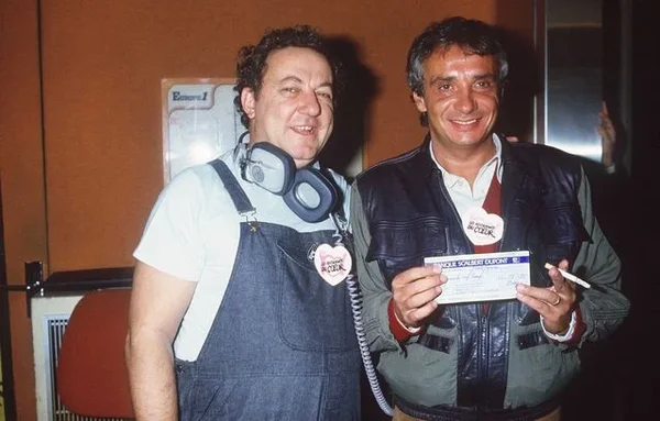 Le comédien Coluche et le chanteur Michel Sardou dévoilant le premier chèque reçu par l'association Restos du cœur dans les locaux d'Europe 1, le 14 décembre 1985.
