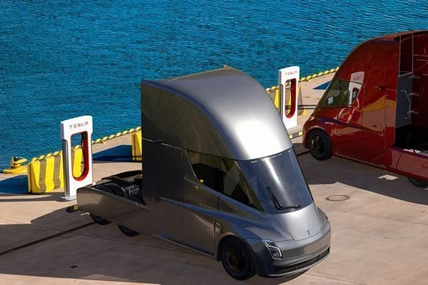 Camion Tesla Semi argenté et camion rouge stationnés à des bornes de recharge Tesla au bord de l'eau, illustration composite en basse résolution.
