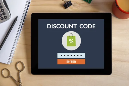 Tablette affichant un code promo 'DISCOUNT CODE' avec un panier et un bouton 'ENTER', posée sur un bureau en bois avec des fournitures de bureau