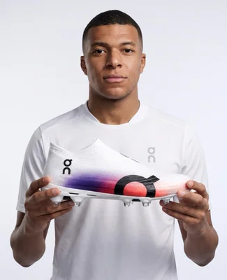Portrait en studio d'un jeune homme ressemblant à Kylian Mbappé, en t-shirt blanc, tenant un crampon de football blanc dégradé de la marque On, sur fond blanc.