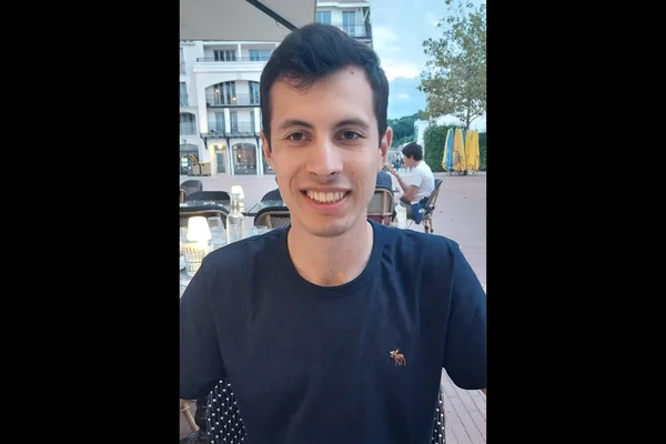 Portrait de Quentin Deranque, jeune militant d'ultra-droite récemment converti au catholicisme, souriant face caméra avec un t-shirt bleu foncé, image encadrée de barres noires.