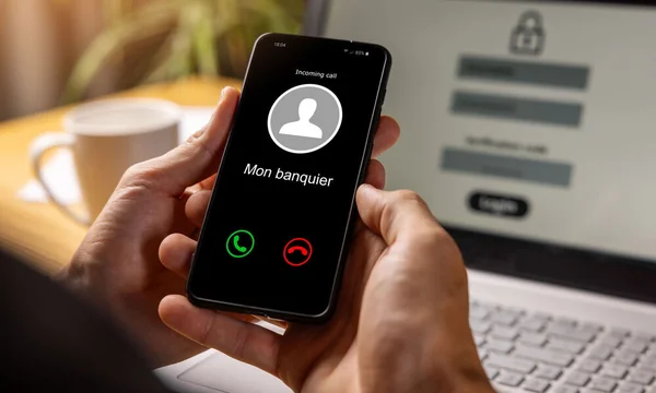 Personne tenant un smartphone affichant un appel entrant de 'Mon banquier' avec un ordinateur portable affichant un écran de connexion en arrière-plan