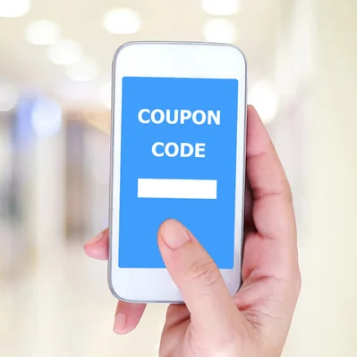 Main tenant un smartphone affichant un code promo 'COUPON CODE' sur fond bleu, dans un environnement intérieur éclairé et flou