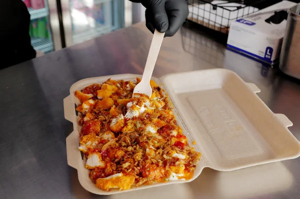 Un conteneur à emporter rempli de Crousty, un plat de riz au poulet frit, tenu par une personne avec un gant noir et une fourchette en plastique blanc dans une cuisine.