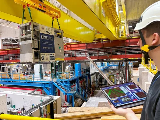Un travailleur en casque blanc et gilet de sécurité tient une tablette affichant des données, tandis qu'un équipement industriel est suspendu par une grue jaune au-dessus d'un plancher métallique dans une installation scientifique.